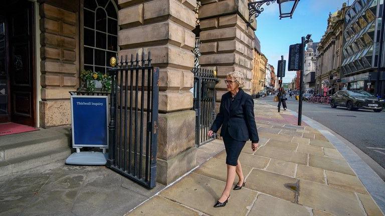 Lady Justice Thirlwall, wearing a black skirt and matching jacket, walks towards the entrance of Liverpool Town Hall, where a blue notice board displays the words 'Thirlwall Inquiry'