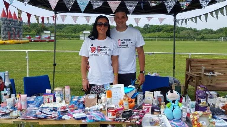 A woman and man stand under a gazebo and behind a table cluttered with various items for sale. They are both wearing white shirts that say "Cardiac Risk in the Young". A field is behind them with a bouncy castle visible on the left.