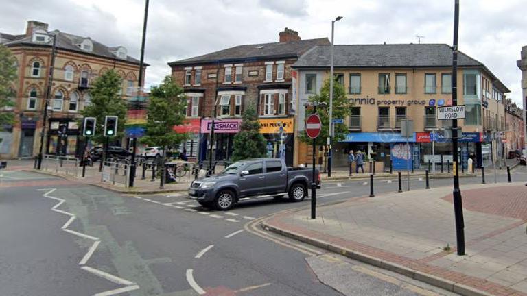 A view of the centre of Withington by the suburb's public library, with a car leaving one road to join another by a set of traffic lights with shops seen in the background. 