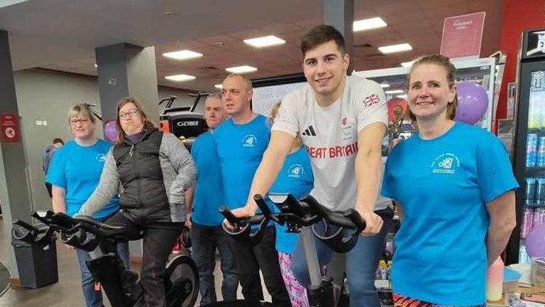 Will Ellard poses on a spin bike wearing a Team GB white T-shirt. BANS volunteers stand next to him and smile at the camera.