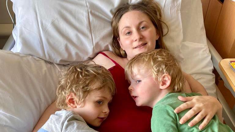 A woman in a bed hugging two children who are lying on top of her.