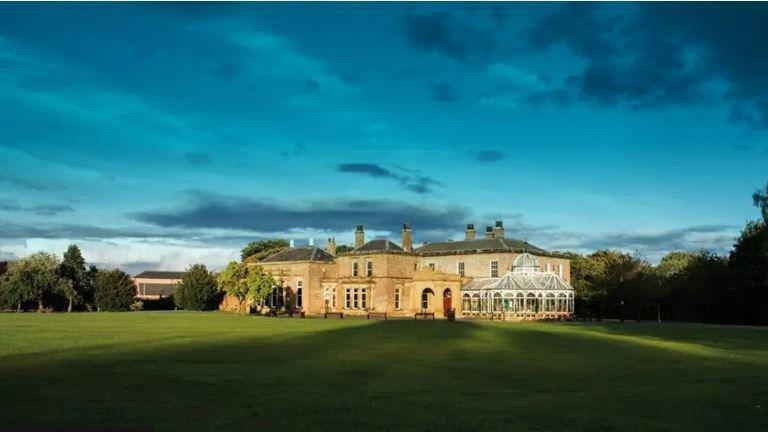 A large stone country manor sits in a expansive green lawn, surrounded by large trees.