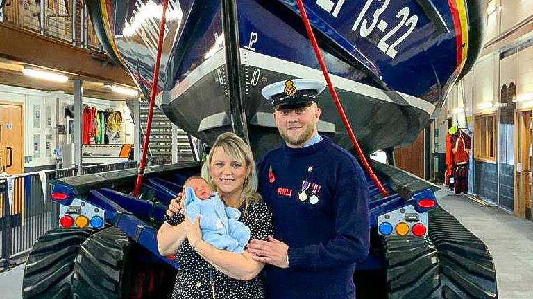 A man in an RNLI navy uniform poses for a photo with his wife and newborn son, in front of a blue lifeboat.