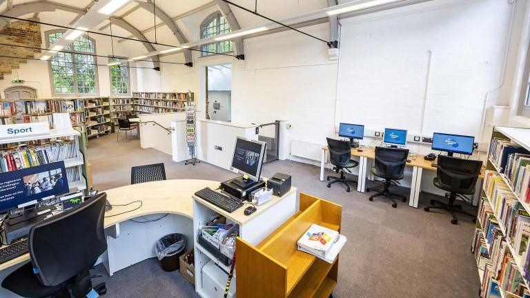 A library room with a row of computer desks, with rows of bookshelves and a large window at one end