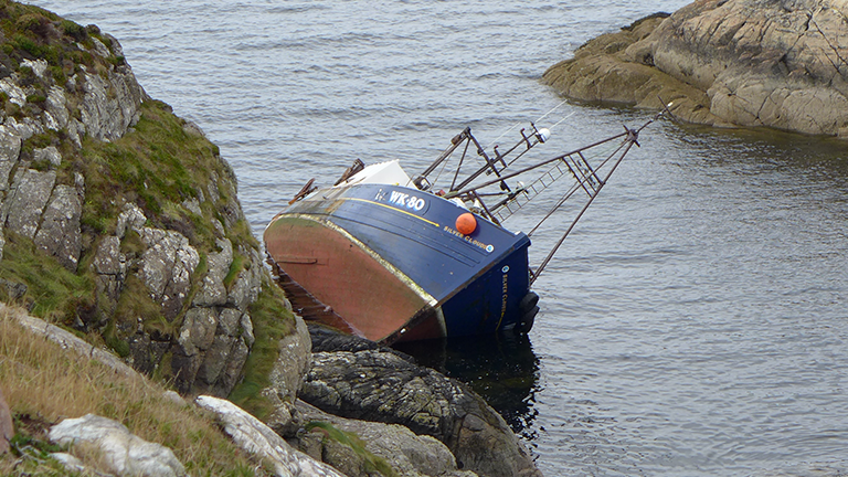 The fishing boat has capsized on a rocky shore. It has a red, white and blue hull and a white cabin.