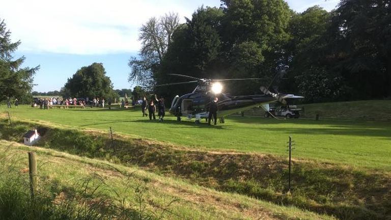 A helicopter on a lawn with a waving man dressed in shorts and a T-shirt boarding