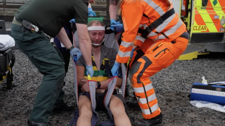 Man with head and body immobilised in a sling being carried towards an ambulance