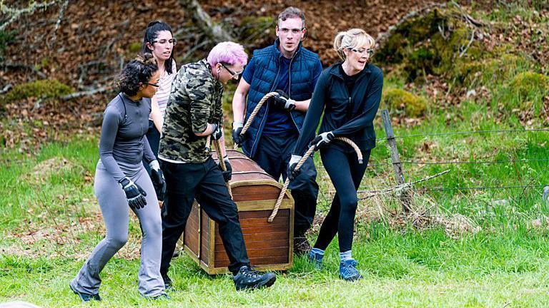 Alex, (centre with pink hair) and four of his fellow contestants, carry a chest in one of the challenges - they are wearing trainers and safety goggles