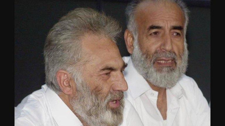 Two men with grey hair and beards, both wearing open-necked white shirts, speak into microphones at a press conference.