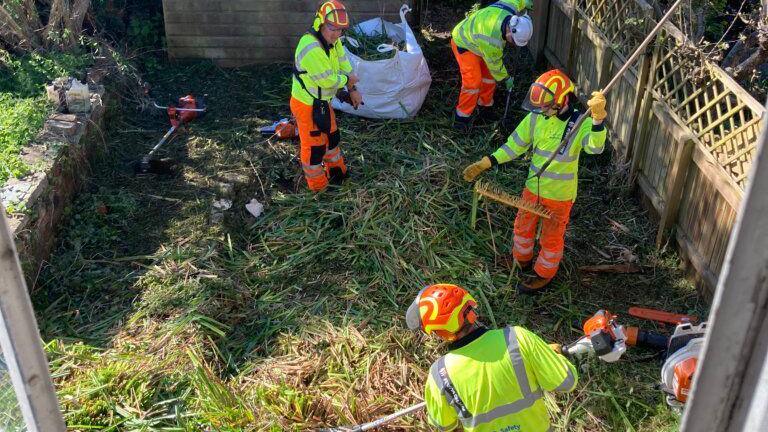 A team from Ringway Infrastructure Services wearing hi-vis clothes helping clear the garden on a sunny day. There is a garden shed in the background.