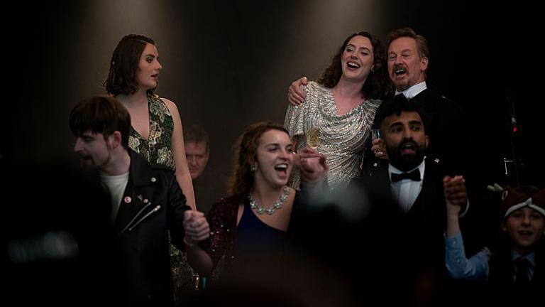 A scene from An t-Eilean (The Island) with the characters Calum (ANDREW MACINNES), Sine (MEREDITH BROOK) Eilidh (SINEAD MACINNES), Douglas (IAIN MACRAE) and Young Ruaraidh (JACOB WATSON) - it appears to be a New Year party, and everyone is singing, laughing or drinking.