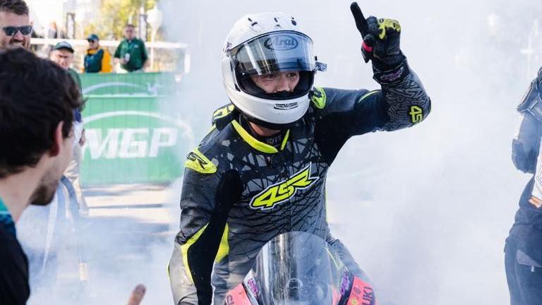Caomahan Canny holding his hand up signalling the number one. He's sitting on his bike with his helmet on after the race and there is smoke around him and a green MGP banner in the background.