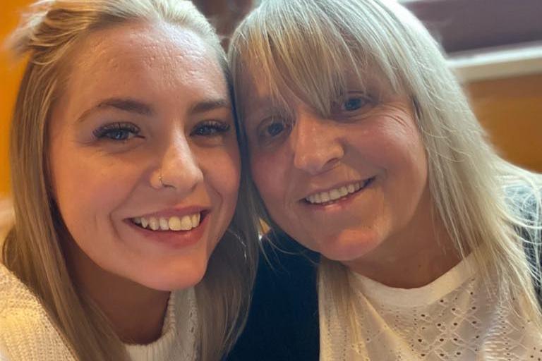 Two women with long blonde hair, both wearing white tops, put their heads together and smile at the camera while the one on the right takes a selfie. Behind them is a yellow wall and a window with a brown frame.