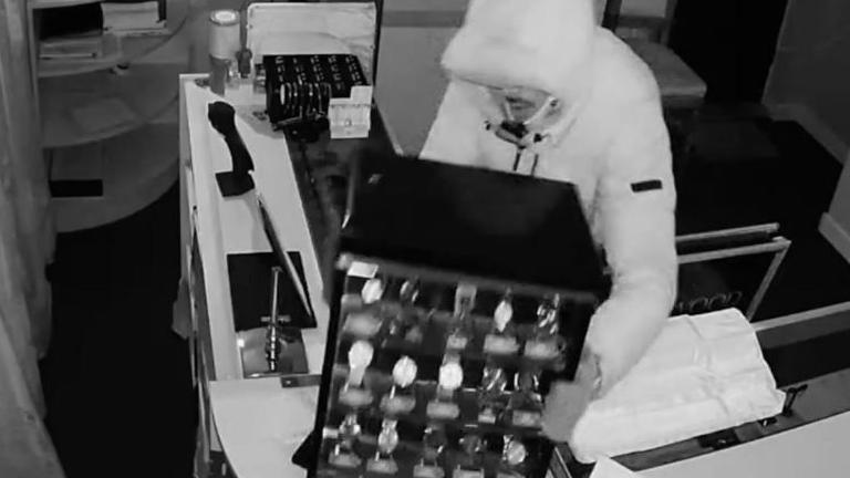 Black and white image of a hooded man picking up a large display of wrist watches from a jewellery counter.