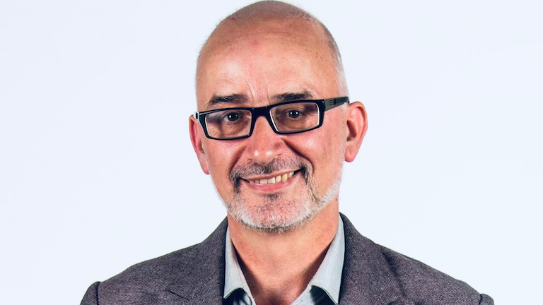 A man wearing black-framed glasses, he has a black and white beared and is smiling. He is wearing a black suit with a grey shirt.