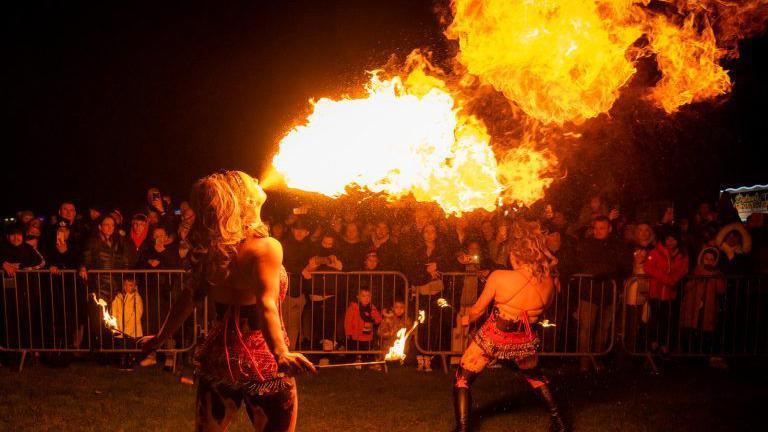 A woman dressed in a cabaret outfit breathes fire into the night sky in front of a crowd of people watching from behind a barricade. She is holding a stick with the end lit with a flame. Another woman, similarly dressed, dances nearby.