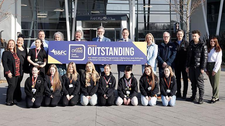 Staff and students pose for a group photograph in the sunshine outside Blackpool and The Fylde College. A large purple sign with the word "OUTSTANDING" in large white writing is held up.