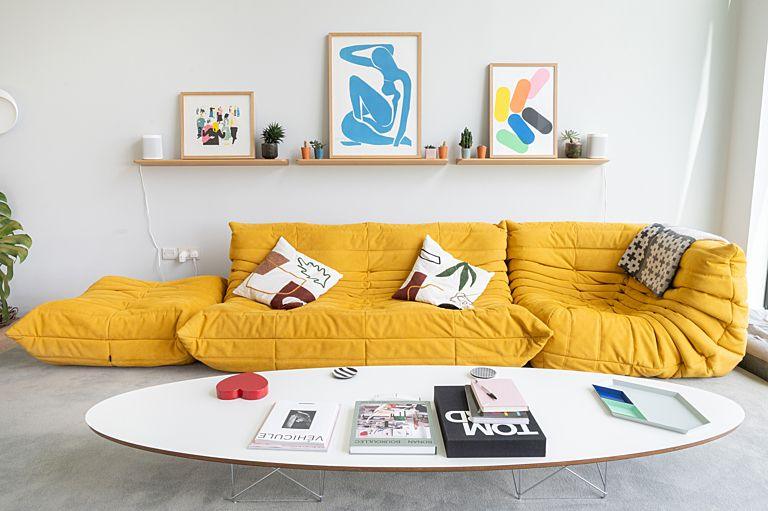 A closer view at the bright yellow sofa which sits against a wall with a picture shelf above, carrying large artworks in bright colours. A stylish white oval coffee table sits in front of it with some style and design books and the red SHOTY heartbeside them.