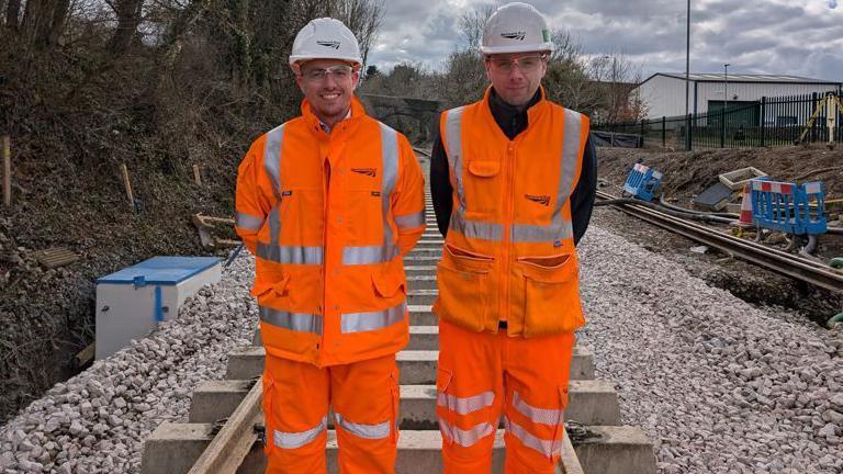 Two men are dressed in orange high vis and hard hats standing on a railway track.