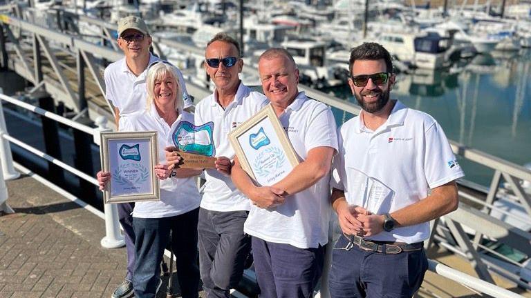 A picture of five people stood leaning on railings in front of the marina.  They are all wearing white polo shirts with a number of people holding framed awards.