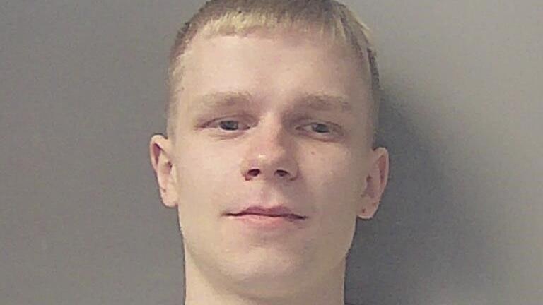 Kairiukstis has short, light-colored hair and is standing against a plain gray background. He is facing forward, and his ears are visible on both sides. He is smiling in the mugshot image.
