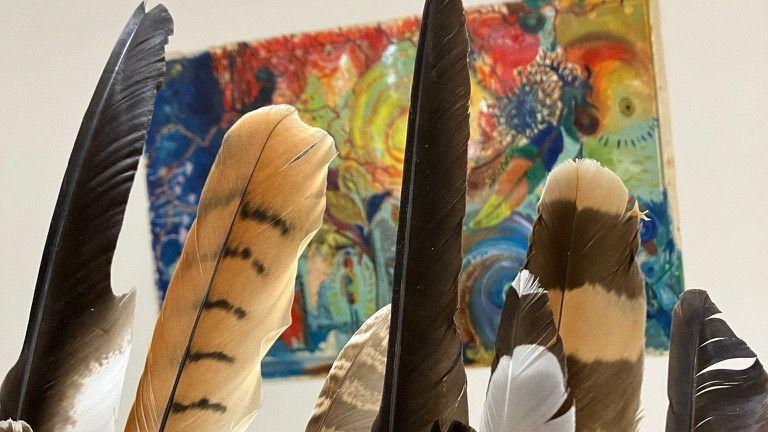 A collection of black, white and black and white feathers all placed in front of a colourful painting inside a room with cream walls.