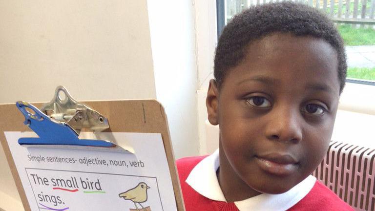 Kwaku Frimpon in school uniform wearing a white t-shirt and red jumper holding a clipboard with sentences on