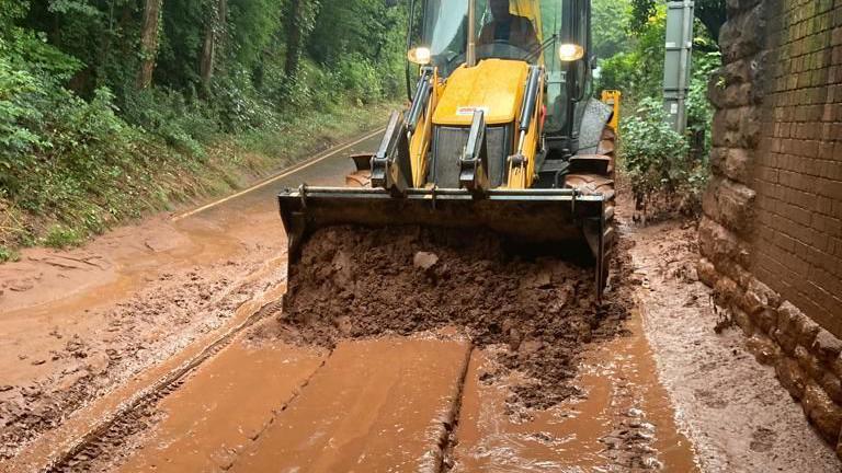 A yellow digger removing thick mud from the road beneath a railway bridge.