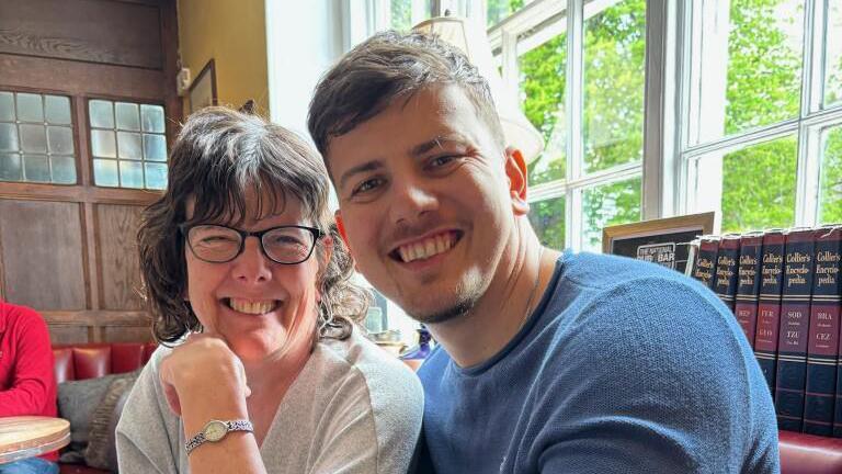 A woman with shoulder length brown hair, black glasses and a white top sits next to a younger man with short dark hair and a beard who is wearing a blue long sleeved top. They are smiling at the camera which is positioned to the side of them.