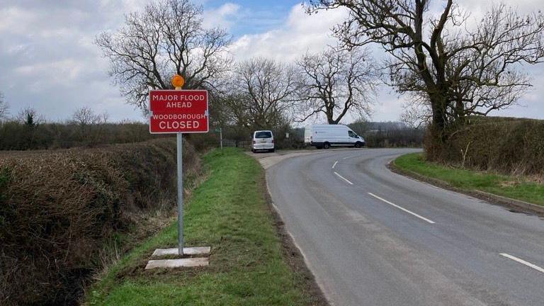 A red warning road sign on a metal pole beside a single track rural road.