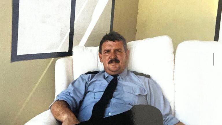 Jeff Lovell sitting on a white sofa in front of a beige wall. He is wearing a blue fire service shirt as part of his uniform, with black epaulettes on his shoulders. He looks to be about middle-age, has dark hair and a full moustache, and is looking at the camera.