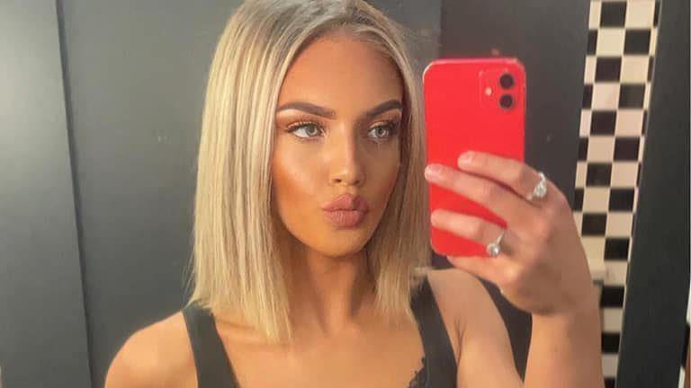 Bradford is taking a photo of herself in a mirror. She has medium length blonde hair and is pouting. Her phone is bright red and the walls behind her are black and white.