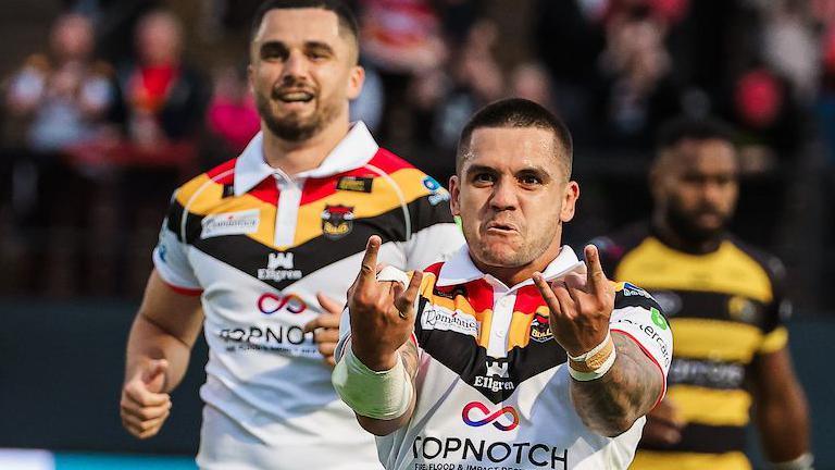 Bradford Bulls player Jayden Nikorima, in white with black, red and gold trim, holds up both hands in the sign of the horns and grimaces to celebrate a try against York Knights, with a team-mate and a York player in the background