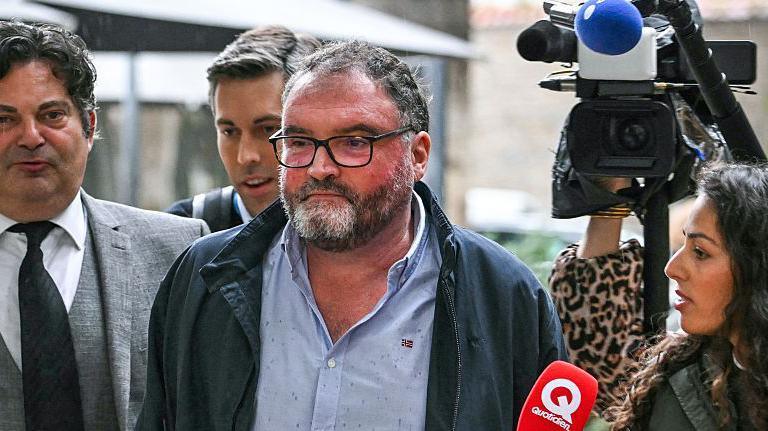 The ex-anaesthetist arrives at court surrounded by reporters wearing a blue anorak and light blue shirt