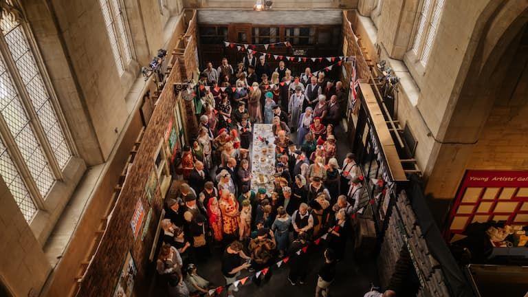 Shot from above depicting a 1940s railway station set created in the chancel gallery at the 20-21 Arts Centre. There are dozens of people in costume looking up towards the camera, along with bunting and people sat at a table eating food.