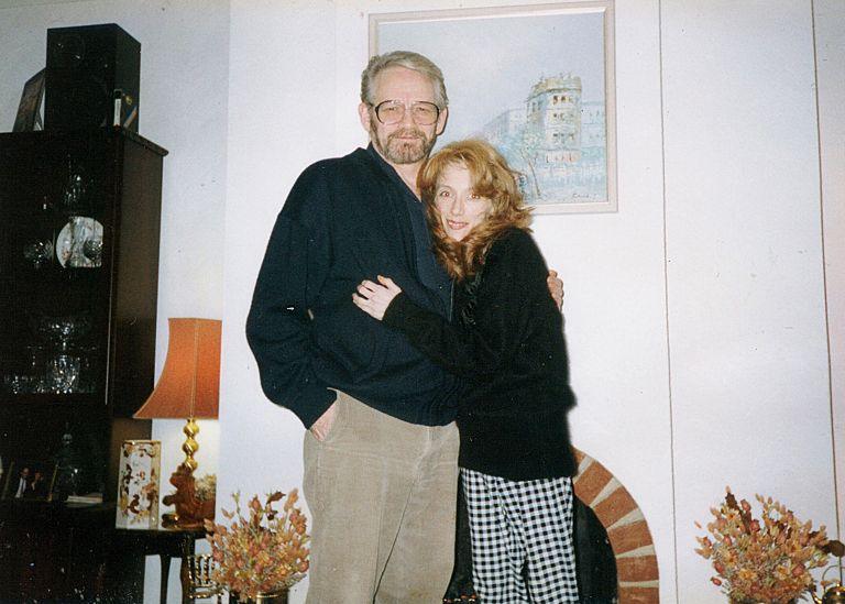 Victor Zavaroni, in beige trousers and a black cardigan, hugs his daughter who is wearing a black jumper and black and white checked trousers. She has long reddish brown hair and her face is drawn. They are in a sitting room beside a fireplace and table lamp.