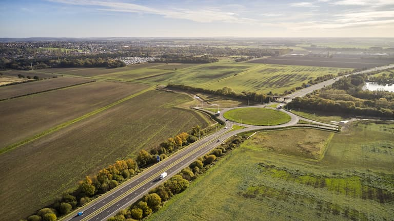 An aerial view of the roadworks on the M181. The road is running through the middle of two large, green fields. A large roundabout is also visible with two unfinished roads leading off it.