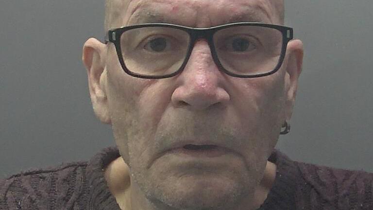 Custody photograph of David Newton, who is clean shaven and bald, looking into the camera, wearing dark glasses, a ring-shaped earring and purple jumper