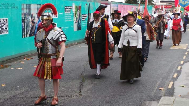 A parade of people in costume - mainly from the English Civil War era, but also with a Roman centurion in front - walking along a street in Gloucester