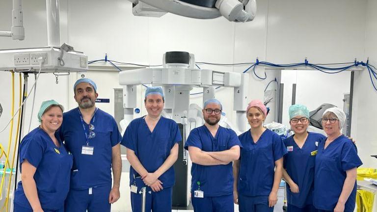 seven people are inside a white operating theatre with white machines in the background. They are all wearing dark blue scrubs and hair nets