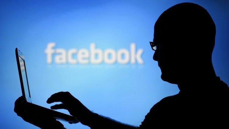 A man with a laptop is silhouetted against a screen with a Facebook logo in the background 14 August 2013