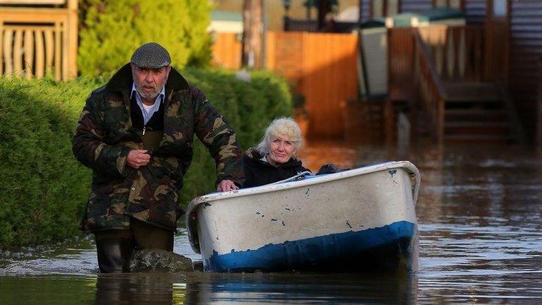 A man wades through water, pulling a boat in which a woman is sitting