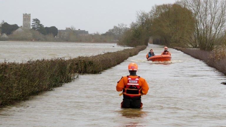 A boat motors along a flooded road towards a rescue worker who stands waist-deep in water