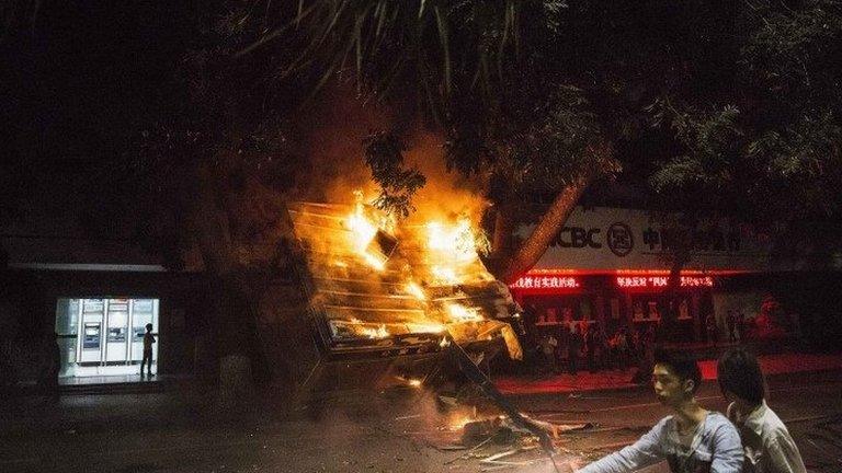 Residents ride past a burning public security kiosk during a protest against a chemical plant project, on a street in Maoming, Guangdong province, 1 April 2014