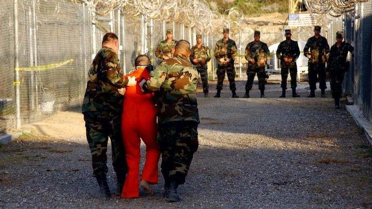 A Guantanamo Bay detainee being walked along by two guards, with more guards looking on