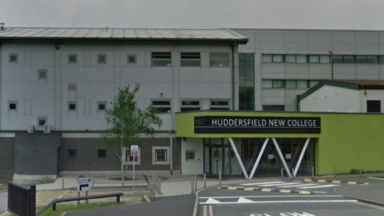 A grey four storey building with a lime green single storey entrance. A sign above the glass doors reads 'Huddersfield New College'. A road with speed bumps curves to the right of the entrance.