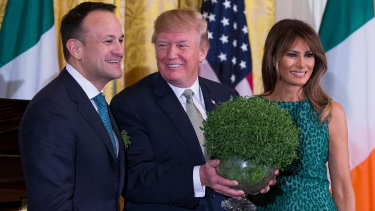 Leo Varadkar smiles on stage beside Donald Trump, who is looking at him and smiling as he holds a large overgrown bowl of shamrocks. Melania Trump is wearing a green patterned dress and is smiling. Two Irish flags and one US flag are on display in the background.