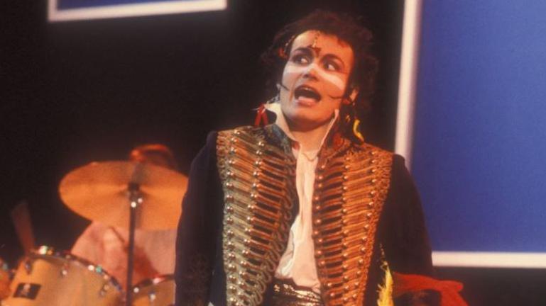 Adam Ant on stage in the 1980s with a drum kit behind him. He is wearing a gold military-style jacket with lots of buttons and brocade and has his traditional white stripe painted across his nose and upper cheeks. He appears to be mid song.