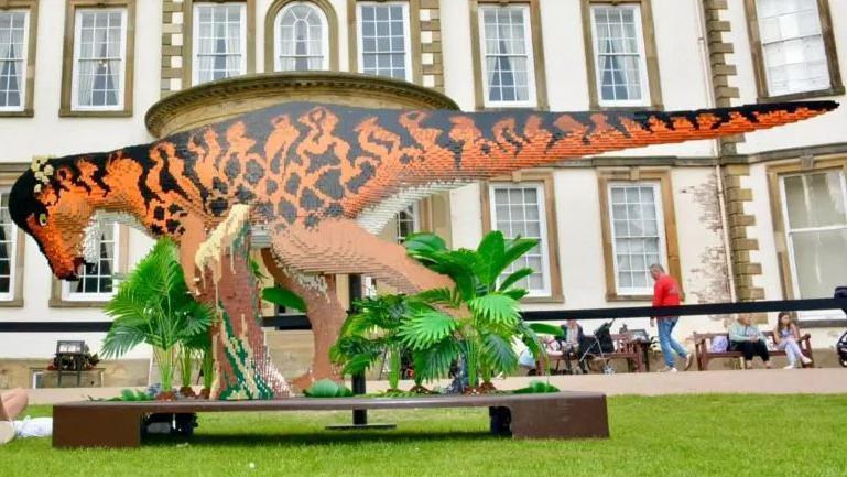 A large brick model of a dinosaur. It is long and is orange and black. 