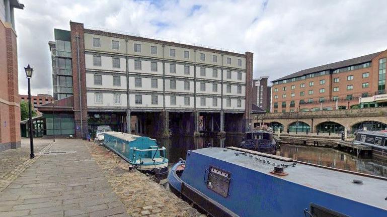 An image of a white painted building which straddles a quayon four columns.  The building is four-storeys high and has ten windows stretching across each level.   in the foreground is the quay with four blue coloured canal boats.  The quayside is a cobbled path with  black traditional iron styled street lights.